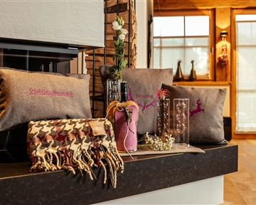 Ein stilvolles Wohnzimmer mit dekorativen Kissen und einer warmen Decke. Im Hintergrund sind große Fenster und eine gemütliche Atmosphäre zu sehen.