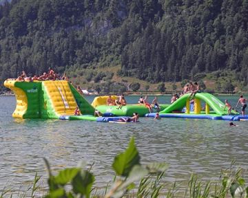 A colorful water play device in the lake with slides and a climbing wall. Many people enjoy the day playing and swimming.