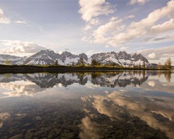 Eine malerische Berglandschaft mit schneebedeckten Gipfeln und einem ruhigen Wasser, das die Wolken spiegelt. Die Szenerie ist friedlich und einladend.