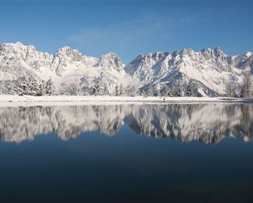 An impressive mountain landscape with snow-capped peaks and a tranquil lake in the foreground. The clear blue sky reflects in the water.