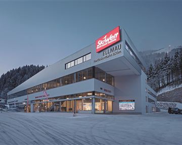 A modern building with large windows and a red sign that shows "Skisport Ellmau." It is located in a snowy environment, surrounded by trees and mountains.