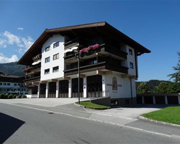 A modern building in alpine style with balconies and flowers. The surroundings are green and sunny, overlooking the mountains.