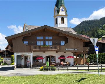 Ein traditionelles alpenländisches Gasthaus mit einem charakteristischen Uhrenturm. Im Vordergrund sind Tische mit roten Schirmen und eine grüne Wiese zu sehen.