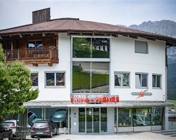 A modern building with an attractive, covered area. In the foreground, some cars and a green environment can be seen.