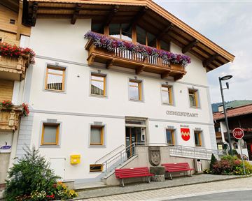 Ein modernes Gebäude des Gemeindeamts mit blühenden Blumenkästen. Vor dem Haus stehen rote Bänke und eine Stoppschild.
