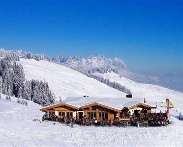 Eine gemütliche Berghütte in den verschneiten Alpen. Umgeben von schneebedeckten Bäumen und strahlend blauem Himmel.
