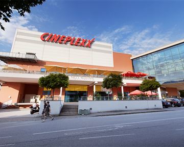 A modern cinema with the inscription "Cineplexx" on the facade. In front of the building, people and plants can be seen.