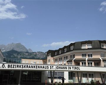 Ein modernes Krankenhausgebäude mit dem Namen "A.O. Bezirkskrankenhaus St. Johann in Tirol". Im Hintergrund sind beeindruckende Berge zu sehen.