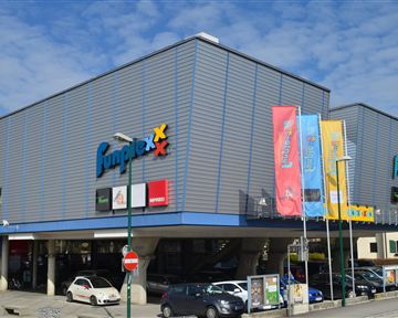 A modern commercial building with the inscription "Jumex". There are several colorful flags and parking spaces in the foreground.