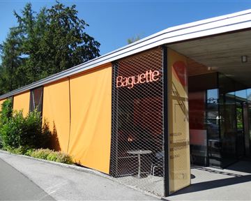 A modern building with a striking orange facade and the inscription "Baguette". Surrounded by green plants and under a clear blue sky.