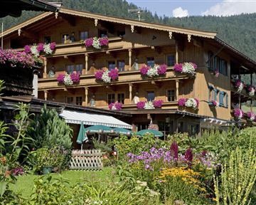 A pretty wooden house with balconies adorned with colorful flowers. In the foreground, there is a beautiful garden with many plants and flowers.