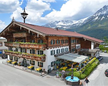 A charming alpine hotel with traditional wooden elements. In the background, impressive mountains and a blue sky can be seen.