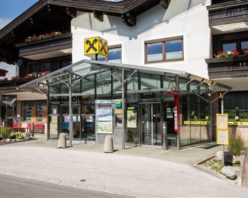 A modern bank with a large glass canopy. It is located on a busy street and is surrounded by well-maintained plants.