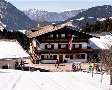 A traditional Alpine house is situated in a snow-covered landscape. Skiers and guests are moving around the area, while the mountains are visible in the background.