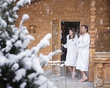 Ein Paar in Bademänteln steht vor einer Holzhütte im Schnee. Dichte Schneeflocken fallen sanft um sie herum.