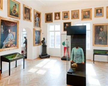 Ein Museum mit historischen Porträts an den Wänden. In der Mitte steht ein Vitrine mit einer Uniform und einem Helm.