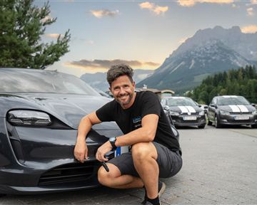 A man is posing smiling next to a modern car in a mountainous landscape. In the background, more cars and a beautiful sky can be seen.