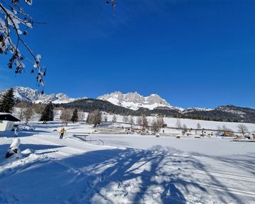 Eine wunderschöne Winterlandschaft mit schneebedeckten Feldern und majestätischen Bergen im Hintergrund. Der Himmel ist klar und blau.