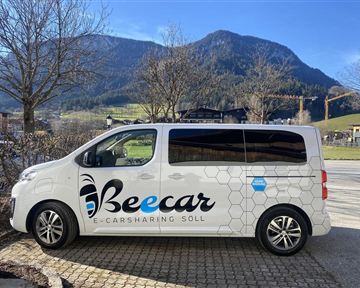 Ein E-Carsharing-Fahrzeug der Marke Beecar steht an einer Straßenkante. Im Hintergrund sind Berge und ein klarer Himmel sichtbar.