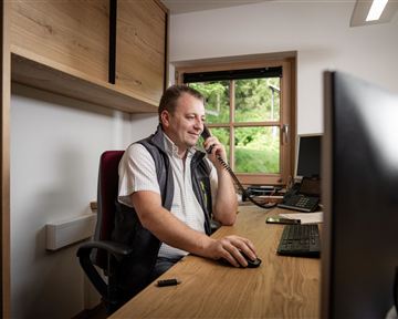 Ein Mann sitzt an einem Schreibtisch und telefoniert. Im Hintergrund ist ein Fenster mit Blick ins Grüne zu sehen.
