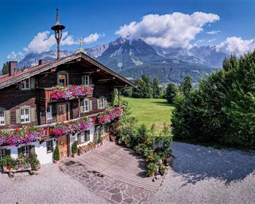 Ein malerisches Bauernhaus mit bunten Blumen und einer idyllischen Umgebung. Im Hintergrund erheben sich majestätische Berge unter einem blauen Himmel.