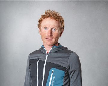 Ein Mann mit roten Haaren trägt eine sportliche Jacke. Er steht vor einem neutralen Hintergrund und schaut direkt in die Kamera.