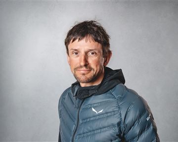 Ein Mann mit kurzer Frisur trägt eine blaue Daunenjacke. Er steht vor einem neutralen, grauen Hintergrund.