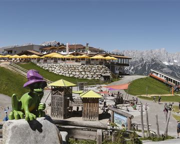 A mountain landscape with a restaurant and sunny terraces. In the foreground stands a green figure with a purple hat and playground equipment for children.