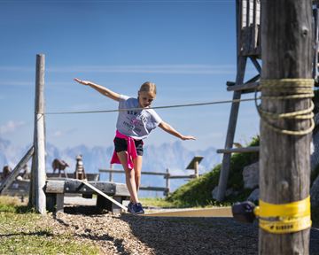 Ein Mädchen balanciert auf einem Seil in den Bergen. Im Hintergrund sind eine schöne Landschaft und eine Holzstruktur sichtbar.