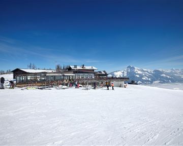A mountainous winter landscape with plenty of snow and a clear blue sky. In the foreground stands a ski lodge, surrounded by skiers.