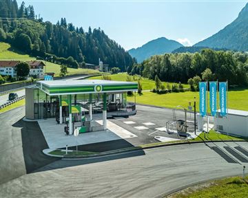 A modern gas station in a picturesque mountain landscape. The surroundings are green with hills and blue mountains in the background.