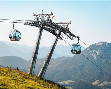 Eine Seilbahn mit blauen Gondeln schwebt über eine grüne Wiese. Im Hintergrund sind majestätische Berge und ein klarer Himmel zu sehen.