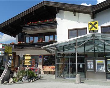 A modern building with a glass facade and a covered entrance. In the background, a traditional Alpine house with flower balconies can be seen.