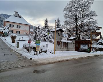 Eine verschneite Straße in einem ruhigen Dorf. Im Hintergrund sind Häuser und verschneite Bäume zu sehen.