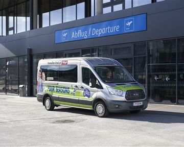 Ein Linienbus steht vor einem modernen Flughafengebäude. Über dem Bus ist das Schild "Abflug / Departure" sichtbar.