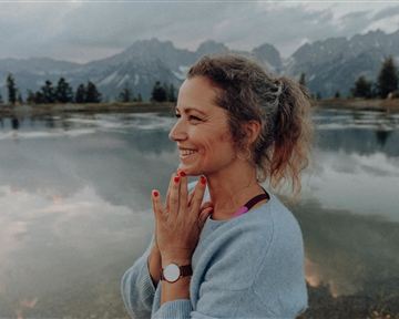 A woman stands at the edge of a calm lake and smiles. In the background, mountains and a cloudy sky are visible.