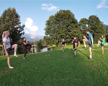 A group of people is exercising on a meadow. Trees and mountains are visible in the background.