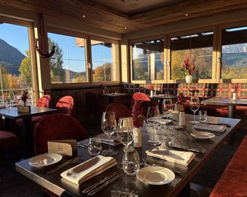Ein elegantes Restaurant mit Holzdekor und roten Stühlen. Der Tisch ist festlich gedeckt mit Gläsern und Geschirr, und große Fenster bieten einen Blick auf die Landschaft.