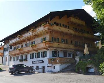 A charming building in Bavarian style with large windows and balconies. In front of the house, there are cars and sun umbrellas on the terrace.
