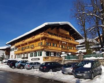 A traditional alpine building with a wooden balcony and green shutters. The surroundings are snow-covered and several cars are parked on the street.