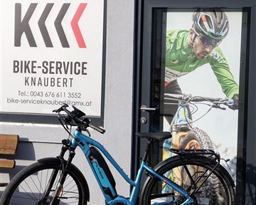 Ein blaues E-Bike steht vor einem Geschäft mit dem Namen "Bike-Service Knaubert". Im Hintergrund ist ein großes Bild eines Radfahrers zu sehen.