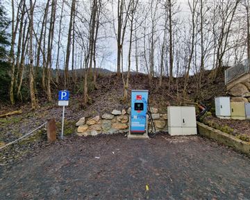 A charging station for electric cars is located in a rural area with trees in the background. Next to the station, there is a parking lot.