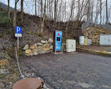 Eine Ladesäule für Elektroautos steht an einem Parkplatz in einer bewaldeten Umgebung. Der Boden ist kiesig und es gibt eine kleine Felswand im Hintergrund.
