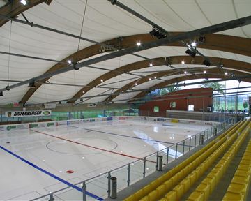Eine moderne Eishalle mit einem leeren Eisfeld und einer geschwungenen Dachkonstruktion. Die Zuschauertribüne ist mit gelben Sitzen ausgestattet.
