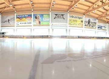 Eine Eishalle mit einer großen, leeren Eisfläche. Der Raum hat hohe Holzdecke und zahlreiche Werbetafeln an den Wänden.