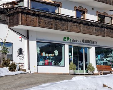 Ein Elektrogeschäft mit großen Fenstern sorgt für einen Blick auf verschiedene Produkte. Vor dem Geschäft liegt Schnee, und die Umgebung ist typisch für eine Berglandschaft.