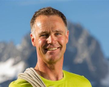 Ein lächelnder Mann trägt ein Seil um die Schulter. Im Hintergrund sind Berge und ein klarer Himmel zu sehen.