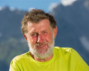 Ein lächelnder Mann mit grauen Haaren und Bart trägt ein leuchtend gelbes T-Shirt. Im Hintergrund sind schöne Berge zu sehen.