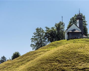 A small church stands on a gentle hill, surrounded by green trees. The sky is clear and blue.