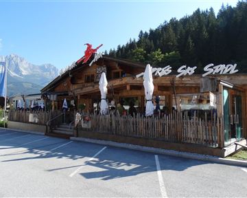 Ein traditionelles Restaurant in einer malerischen Umgebung mit Holzverkleidung und Sonnenschirmen. Im Hintergrund sind Bäume und Berge sichtbar.
